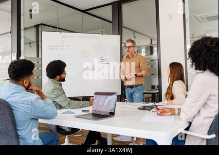 Unternehmensleiter, der eine strategische Präsentation in modernen Büros leitet. Die Teammitglieder hören aktiv zu, engagieren sich für Unternehmensplanung, Marktanalyse und innovative Entscheidungen für den Unternehmenserfolg Stockfoto