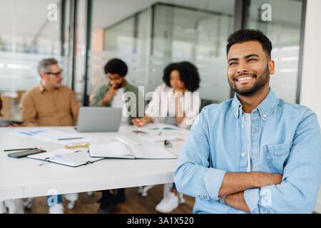 Ein junger Geschäftsmann in lässigem Denim-Hemd sitzt selbstbewusst an einem modernen Arbeitsplatz und strahlt Positivität aus, während Kollegen an einer Teamdiskussion teilnehmen. Stockfoto