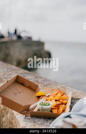 Eine Kiste mit Essen sitzt auf einem Felsvorsprung in der Nähe des Wassers. Die Box enthält Fish and Chips und eine Gabel ist ebenfalls vorhanden. Die Szene hat eine lässige und entspannte Atmosphäre Stockfoto