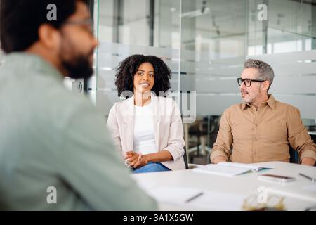 Eine selbstbewusste junge Frau in einem weißen Blazer lächelt, während sie aufmerksam einem Geschäftsgespräch mit ihren Kollegen in einem hellen, professionellen Büro zuhört Stockfoto