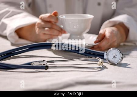Doktor im weißen Kilt, der eine Tasse heißen Kaffee hält. Stethoskop auf einem Tisch im Vordergrund. Stockfoto