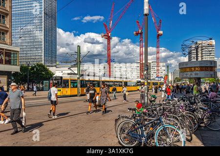 Touristen laufen am Alexanderplatz, Berlin, mit einer gelben Straßenbahn, die vorbeifährt und Baukräne im Hintergrund unter blauem Himmel. Berlin, Deutschland Stockfoto