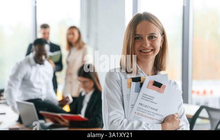 Glückliche Frau steht vor ihren Kollegen. Eine Gruppe von Büroangestellten ist drinnen zusammen. Stockfoto