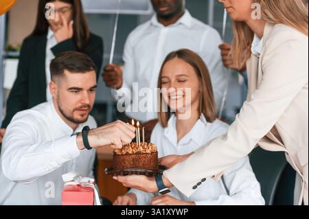 Die Kerzen auf einem Kuchen anzünden. Mitarbeiter, der im Büro Geburtstag hat, Gruppe von Arbeitern. Stockfoto