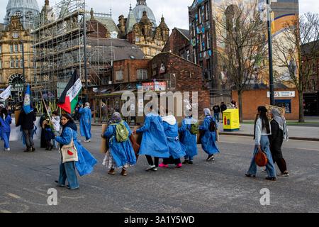 Gruppe marschiert in blauen Mänteln durch eine Stadtstraße, hält Schilder und Fahnen, mit urbanen Gebäuden im Hintergrund an einem bewölkten Tag. Stockfoto