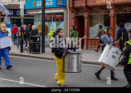Leute, die auf einer Straße marschieren, Trommeln spielen und Schilder halten. Im Hintergrund befinden sich Gebäude und Fußgänger. Stockfoto