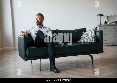 Auf dem Sofa sitzen. Eleganter Mann in Geschäftskleidung ist drinnen mit einem Glas Wasser. Stockfoto