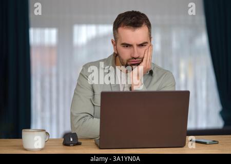 Langweiliger Mann, der Überstunden arbeitete, am Schreibtisch vor einem Laptop saß und verärgert auf den Bildschirm schaute. Stockfoto