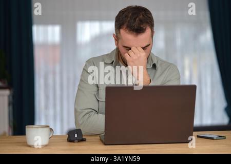 Ein müder Mann arbeitete Überstunden, saß am Schreibtisch vor einem Laptop und reibte sich die Augen mit den Händen. Stockfoto