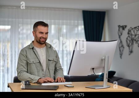 Ein glücklicher Mann, der einen Desktop-Computer benutzt, während er zu Hause arbeitet. Stockfoto