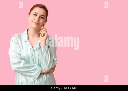 Schöne junge Frau im Seidenschlafanzug mit Gesichtsmassagewerkzeug auf rosa Hintergrund Stockfoto