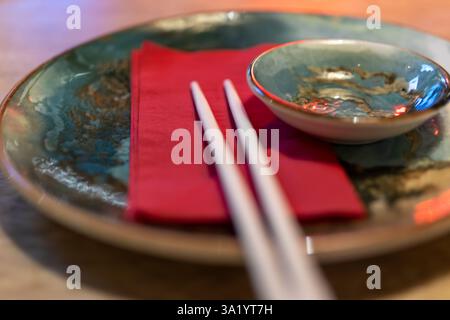 Nahaufnahme eines eleganten asiatischen Tischs mit Essstäbchen, einer roten Serviette und einer Saucenschale auf einem dekorativen Teller. Das Bild erfasst die ess Stockfoto