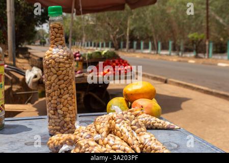 Erdnüsse in Flaschen und frische tropische Früchte werden auf einem Markt am Straßenrand in Enugu, Nigeria, ausgestellt, mit Tomaten und Papayas im Hintergrund Stockfoto