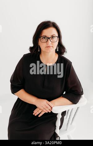Studio-Porträt einer selbstbewussten 40-jährigen Frau, die ein elegantes schwarzes Outfit und eine Brille vor einem sauberen weißen Hintergrund trägt. Stockfoto