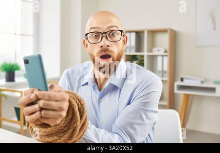 Ein schockierter, besessener Mann, der ein Mobiltelefon sucht und im Büro mit gefesselten Händen am Schreibtisch sitzt. Stockfoto