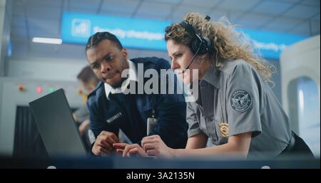 Sicherheitskontrollpunkt am Flughafen: Multiethnische TSA-Beamte im Headset mit Laptop-Computer und Sprechen. Transport Security Administration for Travel Safety. Im Hintergrund arbeiten Flughafenmitarbeiter. Stockfoto