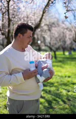 Vater, der sein neugeborenes Baby in der Hand hält, während er eine Milchflasche in einem wunderschönen blühenden Obstgarten hält, und einen friedlichen Moment zusammen genießt Stockfoto