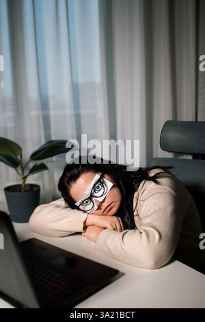 Müde Geschäftsfrau mit gemalten Augen auf Klebenotizen, die Müdigkeit und Kopfschmerzen während der Arbeit im Büro hat. Weibliches Gefühl gestresst und erschöpft von Stockfoto