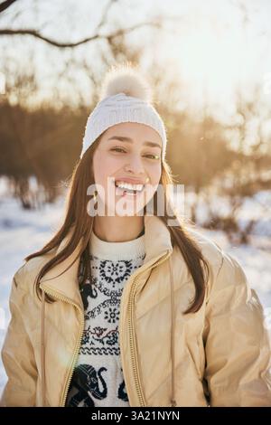 Lächelnde junge Frau in beigefarbener Jacke und Strickmütze genießt Wintersonne mit fröhlichem Ausdruck. Winter Natur. Stockfoto