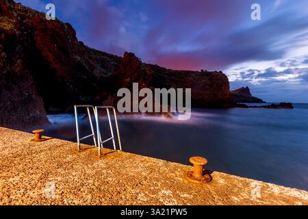 Die Sonne untergeht an der Küste Madeiras und wirft warme Farben über felsige Klippen. Eine verwitterte Leiter führt zu ruhigen Gewässern, die eine ruhige Atmosphäre schaffen Stockfoto