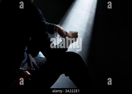 Ein verhafteter Mann sitzt im Dunkeln mit gefesselten Händen und wird von einem Lichtstrahl beleuchtet. Stockfoto