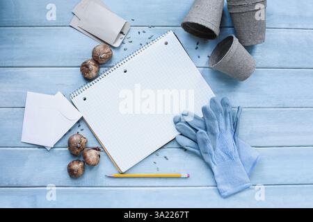 Leeres Gartenzeitschriften-Raster-Notizbuch umgeben von Gartenhandschuhen, Bleistift, Samen, Blumenzwiebeln, Umschlägen und Torftöpfen auf einem rustikalen Holztisch. Tabelle Stockfoto