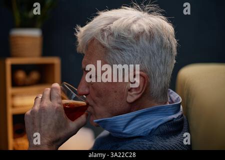 Ein älterer Mann genießt Drinks, während er sich auf einem Stuhl in gemütlicher Umgebung mit sanfter Beleuchtung ausruht und ein ruhiges und zufriedenes Verhalten zeigt Stockfoto