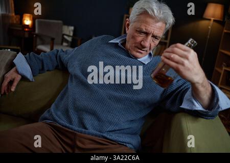 Ein älterer Mann sitzt auf der Couch und untersucht neugierig die Flasche, umgeben von einer gemütlichen Inneneinrichtung und sanfter Beleuchtung. Das Wohnzimmer ist mit Bücherregal, Stuhl und warmer Beleuchtung ausgestattet Stockfoto