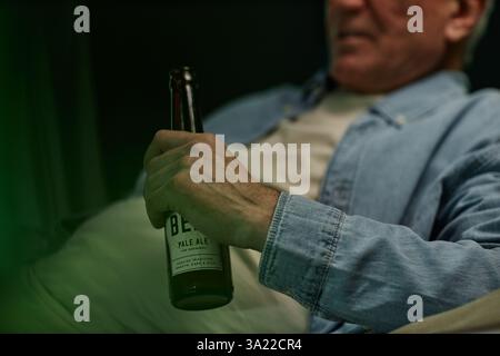 Mann mittleren Alters hält eine Flasche Bier im Sitzen und trägt ein lässiges Jeansshirt. Konzentrieren Sie sich auf Hand und Flasche, mit unscharfem Gesicht, das eine entspannte Atmosphäre vermittelt Stockfoto