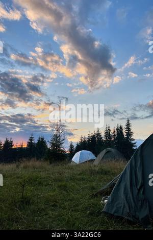 Wenige Touristenzelte in einem grasbewachsenen Feld am Abend, umgeben von Bäumen und Hügeln unter atemberaubendem Sonnenuntergangshimmel. Farbenfrohe Wolken verleihen der Szene eine schöne Note mit leuchtenden Orange- und Violetttönen am Horizont. Stockfoto