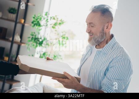 Glücklicher älterer Mann, der eine Pizzabox geliefert bekommt, während er einen ungezwungenen Tag drinnen mit natürlichem Licht in einer gemütlichen Wohnumgebung genießt. Stockfoto