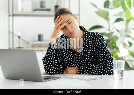 Müde aufgebracht kaukasische Geschäftsfrau sitzt mit geschlossenen Augen an ihrem Schreibtisch im Büro, hält ihren Kopf mit einer Hand frustriert und zeigt Stress und Erschöpfung, während sie an ihrem Laptop arbeitet Stockfoto