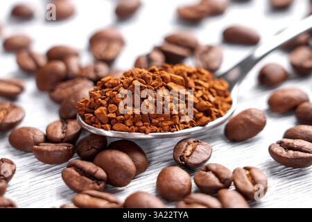 Metalllöffel mit Instant-Kaffeegranulat umgeben von gerösteten Arabica-Bohnen auf weißer Holzoberfläche. Makroaufnahme. Stockfoto