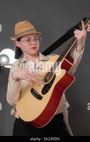 Spielerische Gitarristin, die klassische 6-saitige Akustikgitarre spielt. Freimütige Musikerin mit Brille, gekleidet in schicke Lederjacke und s Stockfoto