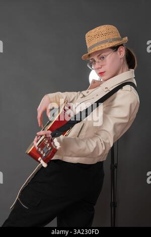 Seitenansicht einer Gitarristin, die 6-saitige Akustikgitarre zupft und in die Kamera schaut. Ernsthafte junge Frau ist eine herausragende Musikerin, trägt Brille und Stockfoto
