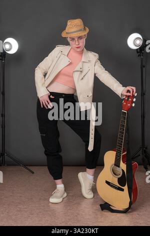Schlanke Gitarristin in voller Länge mit sechs Saiten Akustikgitarre, die selbstbewusst auf der Bühne steht, posiert und in die Kamera blickt. Musikerin in Beige Stockfoto