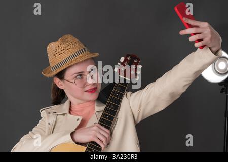 Eine Gitarristin-Influencerin mit Gitarre verwendet Smartphone, schaut sich es an, indem sie Selfie macht, während sie lernt, Gitarre zu spielen, um neue Fotos an ihre Freunde zu schicken und Stockfoto