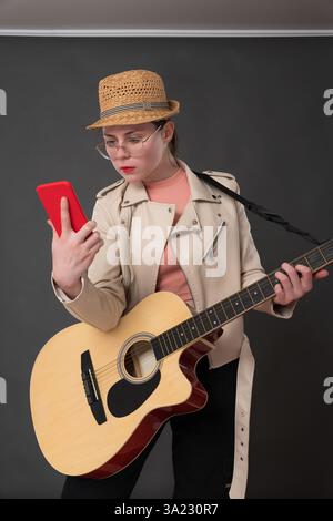 Gitarristin mit Gitarre, die ihr Mobiltelefon benutzt, und dabei neue Akkorde, Melodien oder Songtexte lernt, während sie Gitarre spielt. Musik und m Stockfoto