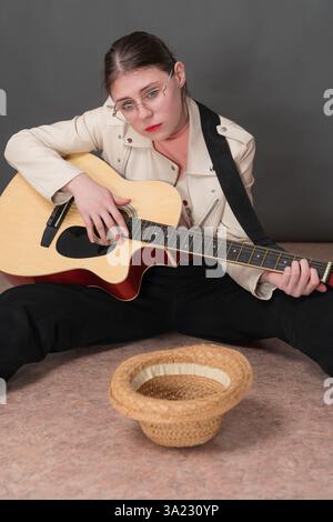 Traurige Gitarristin Straßenmusikerin spielt Akustikgitarre und sieht hoffentlich in die Kamera. Die junge Frau sitzt mit auseinander liegenden Beinen und leerer Strohhut liegt Stockfoto