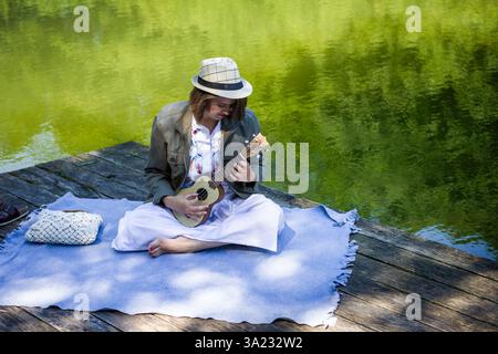 Sorglose junge Frau mit Hut, die Ukulele auf dem Holzpier am See spielt. Genießen Sie Sommerstimmung und lernen Sie Musik. Entspannen Sie sich im Freien Stockfoto