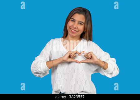 Lächelnde Frau, die Herzzeichen mit Händen auf blauem Hintergrund zeigt und Liebe und Sorgfalt ausdrückt Stockfoto