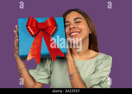 Junge Frau, die eine blaue Geschenkbox mit roter Schleife hält, mit geschlossenen Augen lächelt und einen besonderen Anlass feiert Stockfoto