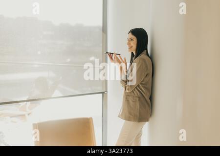Eine junge Frau lehnt sich an eine Wand und lächelt, während sie am Telefon spricht. Sonnenlicht strömt durch das Fenster und hebt ihre natürliche Schönheit und den tran hervor Stockfoto