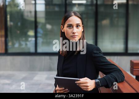 Porträt einer jungen brünetten Geschäftsfrau, die stilvolle schwarze formelle Kleidung trägt. Weibliche Entertainerin, die auf die Kamera blickt, während sie draußen sitzt. Hochformat Stockfoto