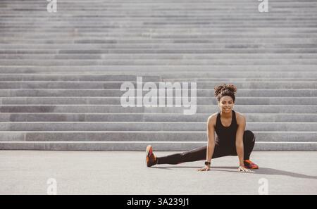 Erschwinglicher Sport im Freien. Lächelnde afroamerikanische Frau in Sportkleidung mit Fitness-Tracker und Kopfhörer tun Stretch für Bein, in der Nähe der Stadt Treppe Stockfoto