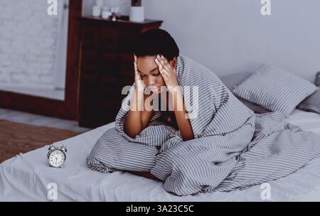 Das afroamerikanische Mädchen in Decke sitzt auf dem Bett mit Wecker Stockfoto