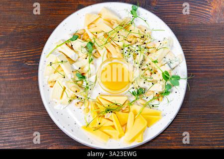 Eine Vielzahl von Käsesorten ist wunderschön auf einer runden Platte angeordnet, begleitet von einer kleinen Schüssel Honig. Frisches Grün verleiht diesem ap einen lebendigen Touch Stockfoto