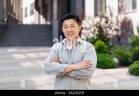 Porträt eines selbstbewussten asiatischen Unternehmers, der mit gekreuzten Armen posiert und die Kamera in der Nähe des Bürogebäudes anschaut Stockfoto