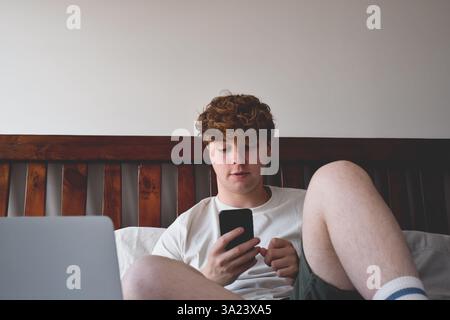 Junger Mann, der mit dem Handy auf dem Bett sitzt Stockfoto