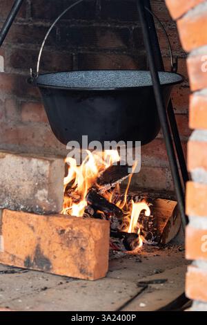 Ein gusseiserner Topf hängt über knisternden Flammen in einem rustikalen Kamin. Das warme Leuchten beleuchtet die Backsteinstruktur und schafft eine gemütliche und einladende Atmosphäre Stockfoto